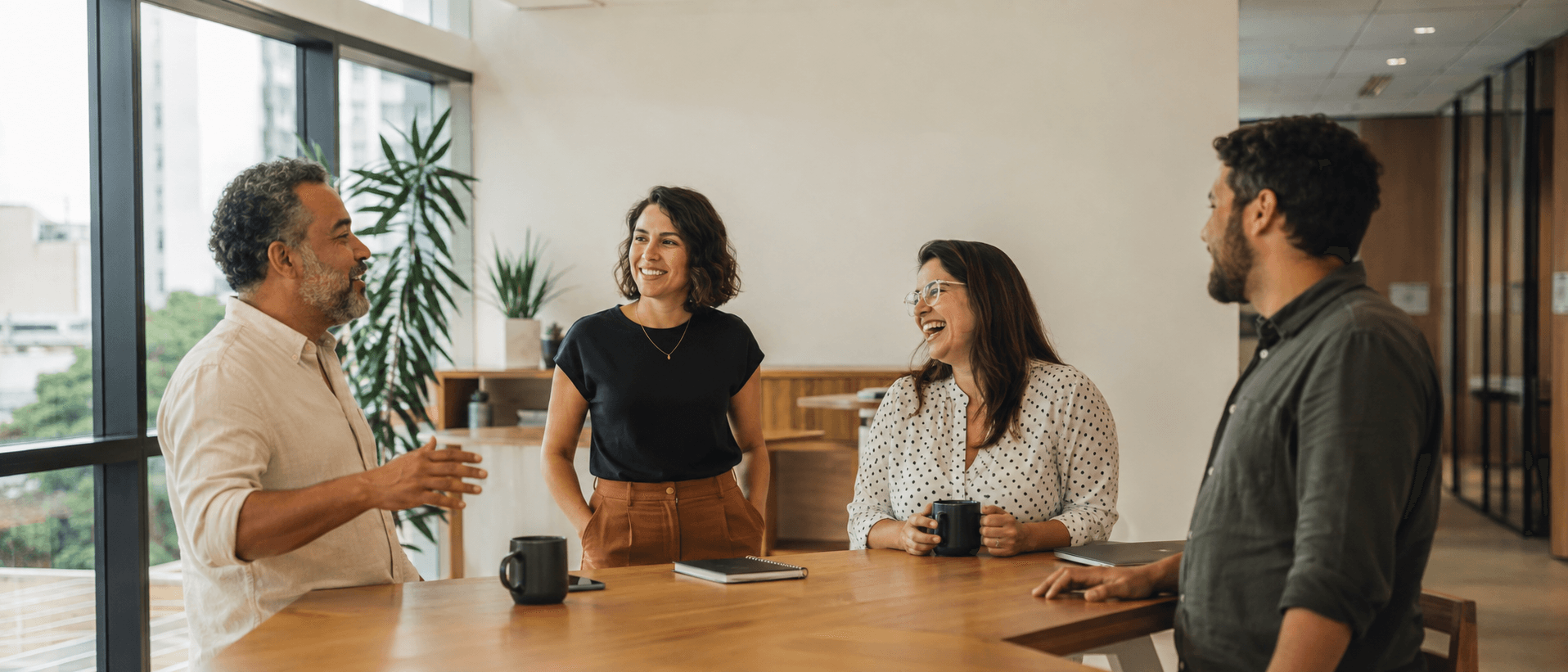 Quatro colegas de trabalho conversando de pé ao redor de uma mesa de madeira, num escritório claro com janela de piso ao teto e plantas