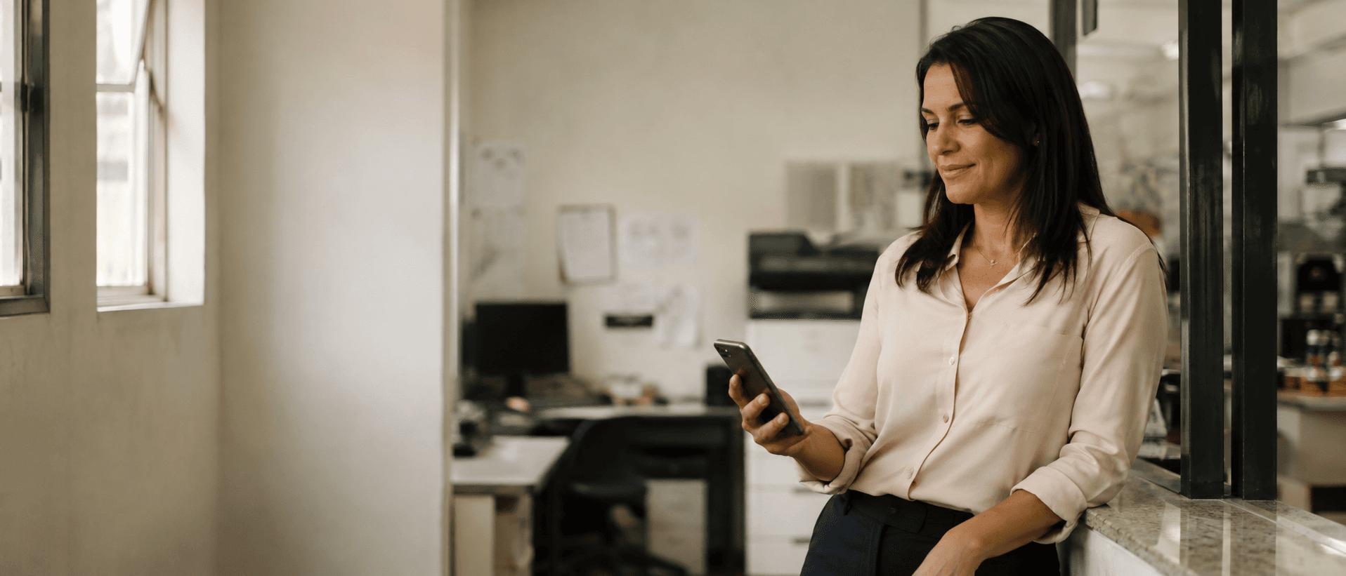 Mulher encostada num balcão, olhando o celular com tranquilidade durante uma pausa, com o próprio posto de trabalho desfocado ao fundo